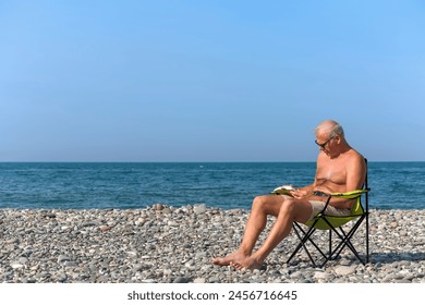 An elderly man is reading a book while sitting in a folding chair on a pebble beach on a hot day