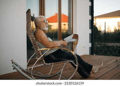 An elderly man relaxes in a rocking chair on a serene porch, enjoying a moment of tranquility in the early evening light - Powered by Shutterstock