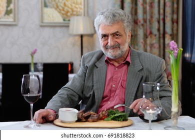 An Elderly Man In The Restaurant Sitting At A Table,eating Meat And Drinking Wine. Interior Photos.