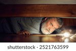 Elderly man with serious and curious face looking under bed with flashlight. Secrets, hidden objects. Hide and seek game. Concept of human emotions, lifestyle, holiday