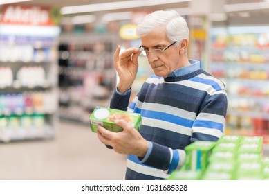 Elderly Man Shopping In Local Supermarket. He Is Holding Box And Reading Nutrition Label.