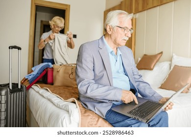 An elderly man sits on a hotel bed using a laptop while his wife packs clothes into a bag. Their luggage is nearby, indicating they are preparing for travel or leaving the hotel. - Powered by Shutterstock