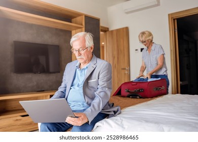 An elderly man sits on a hotel bed using a laptop while his wife packs clothes into a bag. Their luggage is nearby, indicating they are preparing for travel or leaving the hotel. - Powered by Shutterstock