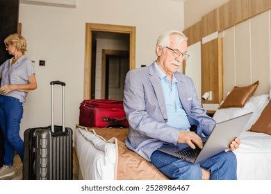 An elderly man sits on a hotel bed using a laptop while his wife packs clothes into a bag. Their luggage is nearby, indicating they are preparing for travel or leaving the hotel. - Powered by Shutterstock