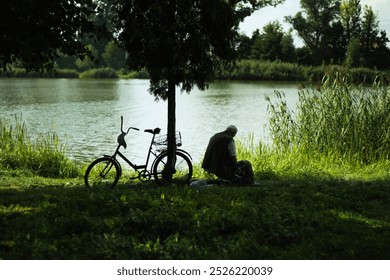 An elderly man sits peacefully by a tranquil lake, surrounded by lush forest greenery. His weathered face reflects a lifetime of stories as he gazes at the stunning view. A vintage bicycle rests nearb - Powered by Shutterstock