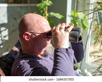 Elderly Man Sittiing, Looking Through A Glass Door With Binoculars As He Watches Something In The Distance