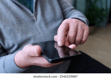 An elderly man is sitting by the window with a smartphone in his hands. The smartphone is in the hands of an elderly man