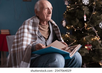 Elderly man sitting in cozy chair, wrapped in checkered blanket, looking through old photo album by decorated Christmas tree. Festive atmosphere with warm holiday lights and nostalgic memories. - Powered by Shutterstock