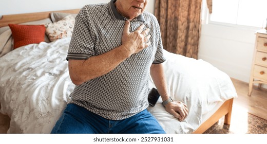 Elderly man sitting on bed, hand on chest, appearing unwell. Casual attire, bedroom setting. Concerned expression, elderly man, health issue, bedroom scene. Elderly man with chest pain, heart attack. - Powered by Shutterstock