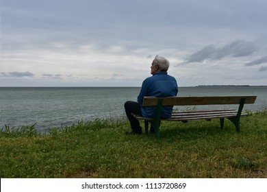 Elderly man sitting on an old wooden bench above the see in a cloudy day, contemplating the nostalgic sea-view  - Powered by Shutterstock