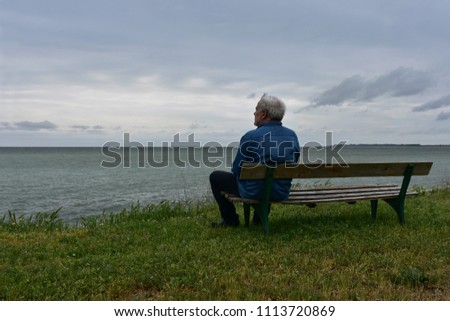 Elderly man sitting on an old wooden bench above the see in a cloudy day, contemplating the nostalgic sea-view 