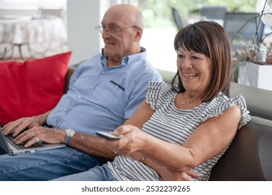 Elderly man sitting on sofa using laptop while wife watches television, two elderly pensioners relaxing in their home with large window overlooking garden - Powered by Shutterstock