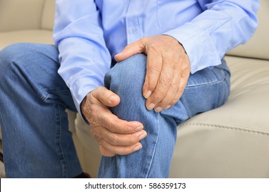 elderly man sitting on a sofa in the living room at home and touching his knee by the pain