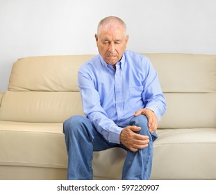 elderly man sitting on a sofa in the living room at home and touching his knee by the pain