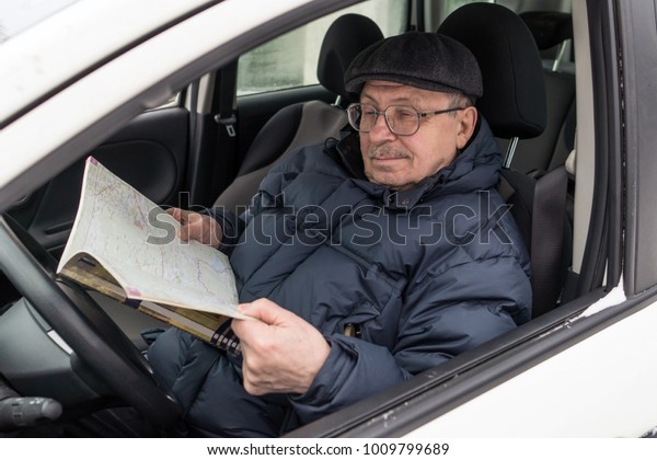 An elderly man is sitting at the wheel of a\
car. The pensioner reviews the road map and chooses a route for the\
trip. Active old age. Winter,\
day.