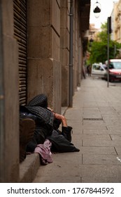 An Elderly Man Is Sleeping On A Doorway By The Metal Shutters Of A Shop During Day Time. He Wears Boots And A Black Coat. Sidewalk, Cars, Trees And Buildings Are Seen In The Background.