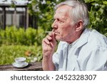 Elderly man smoking a cigarette, wearing a white shirt, outdoors. Reflective, personal moment, suitable for health or lifestyle content.