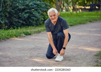 Elderly man in sportive wear ties shoelaces ready for morning jogging or sportive walk stroll in summer park smile look at camera. Healthy active lifestyle of retiree, work out - Powered by Shutterstock