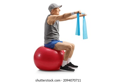 Elderly man in sportswear sitting on a fitness ball and exercising with an elastic band isolated on white background