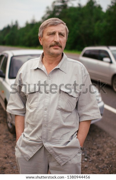 An elderly man stands near his car on the road.\
The old man travels by auto on the highway. senior man standing and\
looking into the distance