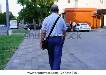 An elderly man strolls down a clean city sidewalk during a warm summer evening. He carries a bag over his shoulder and passes by various shops and parked vehicles.