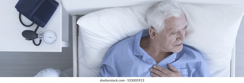 Elderly Man Suffering From Chest Pain Lying In Hospital Bed With Pressure Gauge On A Nightstand