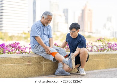 Elderly man suffering knee pain during jogging exercise with his son at park in the city. Adult man gives first aid to father with knee injury. Family relationship and older people health care concept - Powered by Shutterstock