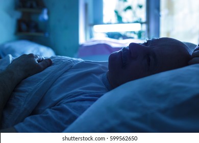 An Elderly Man Suffers From Insomnia, Trying To Sleep. Backlight Image In Blue Tones. 