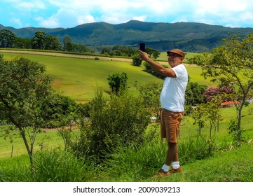 Elderly Man Taking A Selfie In A Rural Town Surrounded By Stunning Nature. Retired Old Man Standing In A Beautiful Green Field Taking A Selfie Around A Beautiful Mountain Landscape.