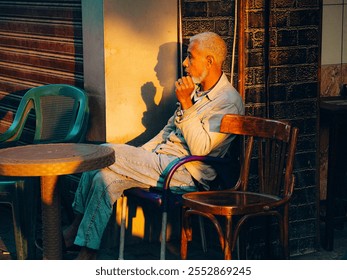 Elderly man in thoughtful pose near wall, casting shadow, surrounded by empty chairs, with warm evening light. - Powered by Shutterstock