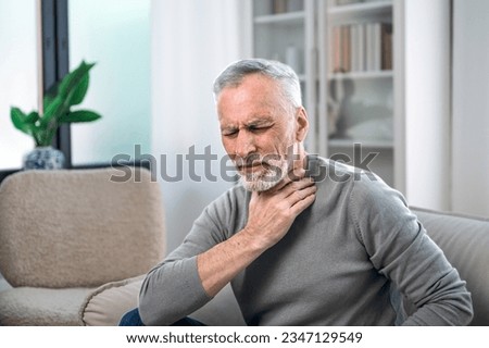 elderly man with throat sore, feeling sick and touching lymph nodes while sitting on couch. male with virus infection stay at home. healthcare concept