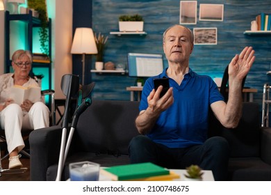 Elderly Man Using Smartphone For Video Call Communication In Living Room. Aged Person Waving At Conference Camera For Online Remote Meeting. Woman Sitting In Wheelchair In Background