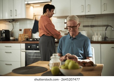 Elderly man using smartphone while sitting at kitchen table, with woman cooking in background, capturing daily life at home, equipped with modern appliances - Powered by Shutterstock - Get 15% off with code: PIKWIZARD15