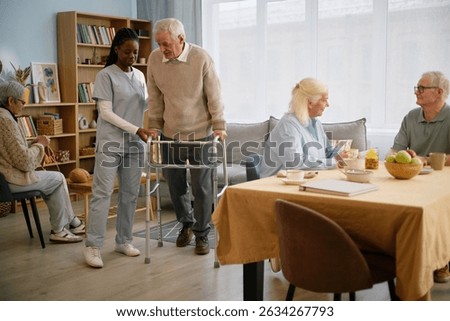 Elderly man using walker assisted by caregiver in care facility with other seniors in background. Room filled with books, chairs, and a table with breakfast items arranged
