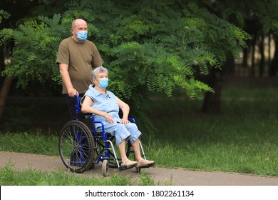 Elderly Man Walking With Disabled Elderly Woman Sitting In Wheelchair Outdoors Wearing Medical Masks