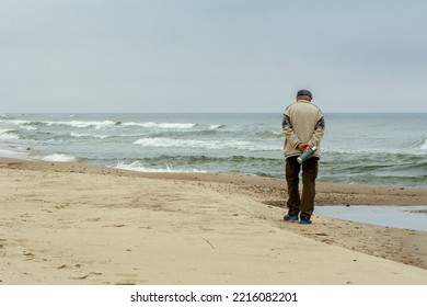 Elderly Man Walking On The Shore Of A Sandy Beach With Beautiful Rough Grey Sea With Waves, Dramatic Cloudy Grey Sky In Winter