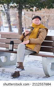 Elderly Man In Warm Coat, Scarf And Cap Having Hot Tea From Thermos While Sitting On Bench In Park On Winter Day