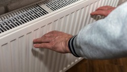 An Elderly Man Warms His Hands Near A Heating Radiator, A Man Froze, A Man Warms Himself Near A Fireplace. A Man Warms Himself Up By An Electric Battery
