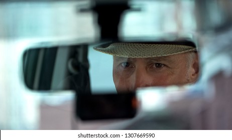 Elderly man wearing hat and looking in rear view car mirror, driver close-up - Powered by Shutterstock