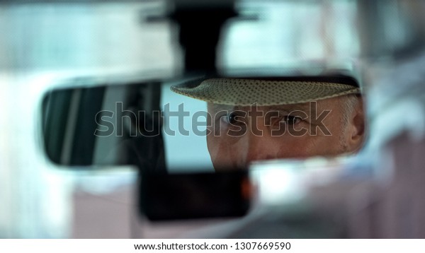 Elderly man wearing hat and looking in rear view\
car mirror, driver\
close-up