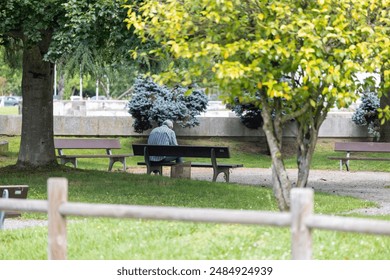 An elderly man, wearing a striped shirt and blue shoes, is seen sitting alone on a park bench in a serene garden setting with trees and a basket nearby. - Powered by Shutterstock