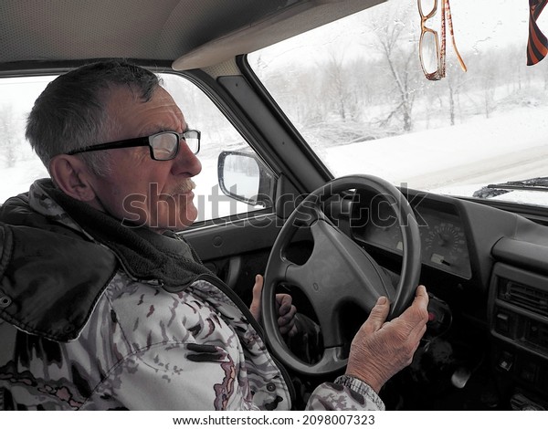 An elderly\
man in a winter jacket driving a Lada car goes fishing in winter.\
Winter and snowfall behind the\
glass.