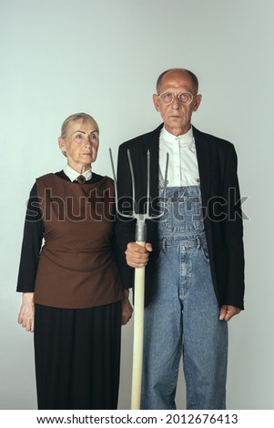 Elderly man and woman in art performance, replica of painting american gothic. Retro style, comparison of eras and cultural concept. Old actor and actress like farmers. Copy space for ad.