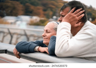 Elderly man and woman are leaning on the railing of the bridge, talking and looking at the city landscape, the old married couple happily spends together, taking care and supporting each other. - Powered by Shutterstock