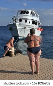 An Elderly Man And Woman With Significant Overweight Are Photographed By A Moored Pleasure Boat On The Pier Of Hurghada. Sunny Winter Day, Egypt.