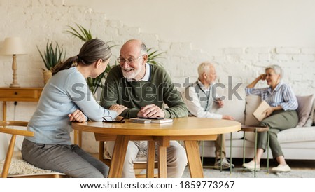 Elderly man and woman sitting at table and enjoying joyful talk, another senior couple communicating in background sitting on sofa in common room of assisted living home