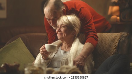 Elderly Man Wrapping Warm Knitted Scarf Around Aged Woman Sitting On Sofa And Drinking Tea Then Hugging Wife In Living Room At Home