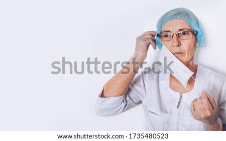 Elderly mature woman doctor or nurse in a white medical coat, gloves, puts on face mask wearing personal protective equiment isolated. Healthcare and medicine concept. Covid-19 pandemic crisis
