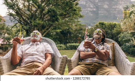 Elderly men with facial mask and cucumber slices on their face at spa. Retired friends enjoying facial spa treatment with juice glasses in hand.