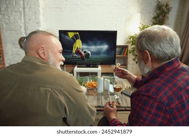 Elderly men, sport fans sitting o couch at home in living room, watching online football match on TV, drinking beer and support favorite team. Concept of sport, championship, leisure and entertainment - Powered by Shutterstock